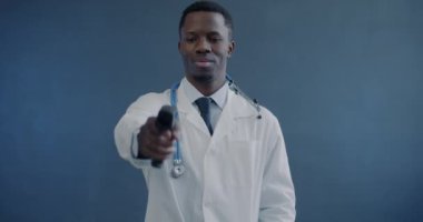 Afrikalı Amerikalı doktorun portresi kameraya bakıyor ve sıcaklığı gri arka planda modern kızılötesi termometreyle kontrol ediyor.