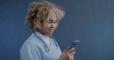 Sosyal medyada mavi arka planda akıllı telefon mesajları kullanan Afrikalı Amerikalı hemşirenin portresi. Tıp ve cihaz kavramı.