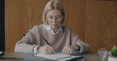 Düşünceli, olgun kadın terapist, modern ofiste masa başında kameraya bakarak yazıyor. Profesyonel meslek ve terapi oturumu kavramı.