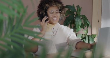 Yaratıcı Afrikalı Amerikalı kadın cep telefonuyla konuşuyor ve güneşli bir günde modern ofiste çalışırken dizüstü bilgisayar kullanıyor. İş iletişimi ve insan kavramı.