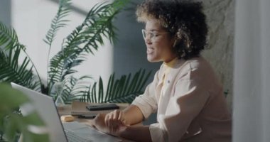 Neşeli genç iş kadını bilgisayarla video görüşmesi yapıyor ve kulaklıkla ofiste iş hakkında konuşuyor. İnternet iletişimi ve girişimci konsepti.