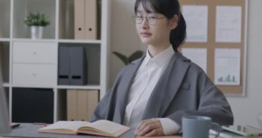 Asyalı barışçıl iş kadınının yavaş çekimde çekilmiş bir portresi. Masasında oturmuş kitap kameraya dönüyor ve ofiste gülümsüyor. Halk ve iş kavramı.
