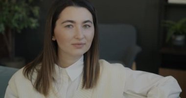 Evdeki koltukta oturan ve kameraya gülümseyen genç bir kadının yavaş çekim portresi. İnsanlar ve olumlu duygu kavramı.