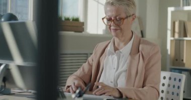 Bilgisayarla çalışan olgun bir iş kadınının yavaş çekimde kameraya dönüp ofiste gülümsemesi. Halk ve iş kavramı.
