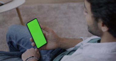Arap adam krom anahtar yeşil ekranlı akıllı telefona bakıyor evde aletle dinlenmekten zevk alıyor. İnsanlar ve aygıt kavramı.