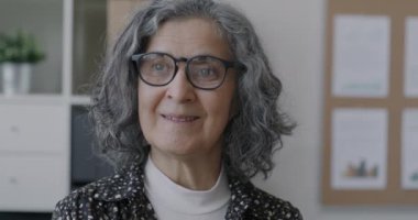 Ofiste kameraya doğru gülümseyen yaşlı iş kadınının yavaş çekim portresi. Deneyimli çalışan ve kariyer kavramı.