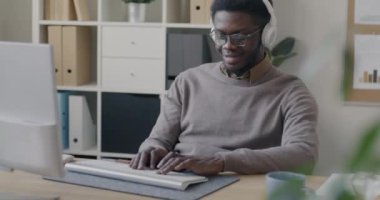 Afrika kökenli Amerikalı bir çalışan bilgisayarla çalışıyor ve ofiste kulaklıkla müzik dinliyor. Modern teknoloji ve işyeri kavramı.