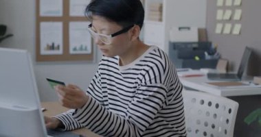 Asyalı iş kadını plastik kredi kartı kullanıyor ve ofiste finans odaklı bilgisayarla yazı yazıyor. İş kadını ve kârlı girişimcilik kavramı.