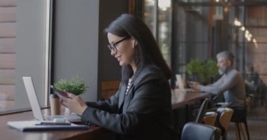 Laptop ve akıllı telefonla çalışan neşeli iş kadını kafedeki masada kablosuz kulaklık kullanıyor. Modern teknoloji ve iş kadını kavramı.