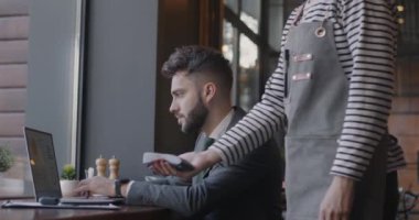 Dizüstü bilgisayarla çalışan genç bir işadamı sonra da kafeteryada garsonla konuşan bir akıllı telefonla ödeme yapıyor. Modern teknoloji ve iş adamı kavramı.