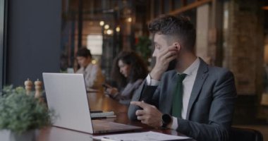 Kablosuz kulaklık takan genç işadamı kafede laptopla konuşuyor ve kahve içiyor. Modern teknoloji ve iş iletişimi kavramı.