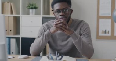 Ofiste oturan ve ciddi bir yüzle kameraya bakan dalgın Afro-Amerikan iş adamının yavaş çekim portresi. İş ve insan kavramı.