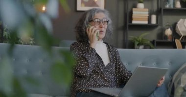 Cep telefonuyla konuşan ve dizüstü bilgisayarı tutan mutlu yaşlı bayan evde başparmak işareti gösteriyor. İletişim ve insan kavramı.