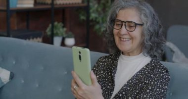 Neşeli, son sınıf öğrencisi bir kadın evde akıllı telefonuyla internetten arama yaparken baş parmak hareketi yapıp gülüyor. İletişim ve emekli kişi kavramı.