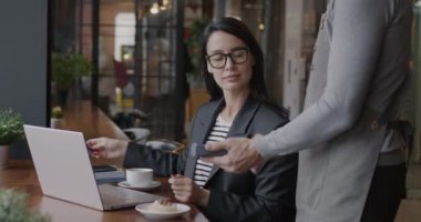 Neşeli genç iş kadını kredi kartıyla ödeme yapıyor ve öğle arasında kafede erkek garsona gülümsüyor. Modern bankacılık teknolojisi ve iş adamı konsepti.