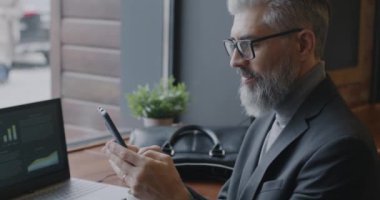 Kafenin içinde sosyal medyada akıllı telefon mesajları kullanan neşeli, olgun bir iş adamı. İletişim ve iş adamı kavramı.