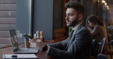 Genç işadamı cep telefonundan dizüstü bilgisayar kullanıyor ve kafede çalışan iş belgelerini kontrol ediyor. Finans ve iletişim kavramı.