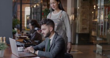 Tasasız genç işadamı laptopla çalışırken ve kafede garsonla konuşurken akıllı saatle ödeme yapıyor. Modern yaşam tarzı ve iş konsepti.