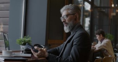 Olgun iş adamı cep telefonuyla konuşuyor, kablosuz kulaklık kullanıyor ve kafede dizüstü bilgisayarla çalışıyor. İş iletişimi ve modern teknoloji kavramı.
