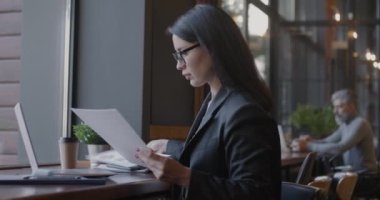 Belgelerle çalışan ve kafede dizüstü bilgisayar kullanan kendine güvenen genç bir iş kadını. Yönetici konsepti için iş belgeleri ve evrak işleri.