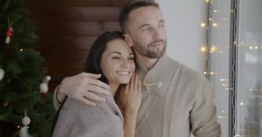 Mutlu adam ve kadın kucaklaşıyorlar ve pencereden dışarı bakıyorlar evde Noel gecesinin tadını çıkarıyorlar. Romantik ilişki ve tatil kutlaması konsepti.