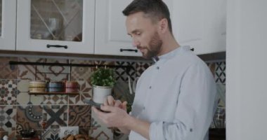 Akıllı telefon kullanan genç adam mutfaktaki sosyal medyada ekran mesajlarına dokunuyor. Modern teknoloji ve iletişim konsepti.