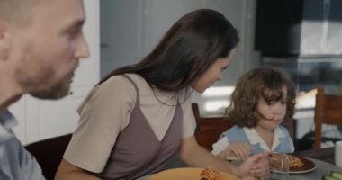 Genç bir kadın, evde kocası ve oğluyla yemek yerken sevimli çocukla konuşuyor. Aile hayatı ve mutlu çocukluk kavramı.