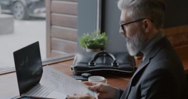 Olgun iş adamı dokümanları okuyor ve kafeteryada kahve içen dizüstü bilgisayarlara bakıyor. Modern teknoloji ve profesyonel işgal kavramı.