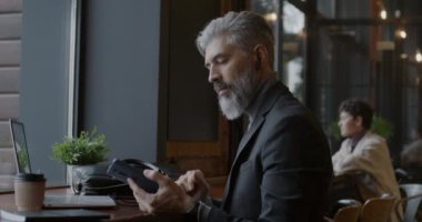 Takım elbiseli deneyimli bir işadamı cep telefonuna cevap verirken kablosuz kulaklık kullanıyor ve kafede dizüstü bilgisayarla çalışıyor. İş iletişimi ve kişi kavramı.