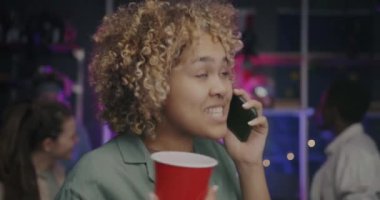 Neşeli Afrikalı Amerikalı kadın cep telefonuyla konuşuyor ve içkiyle dans ediyor. Partide arkadaşlarıyla eğleniyor. Gençlik ve modern teknoloji kavramı.