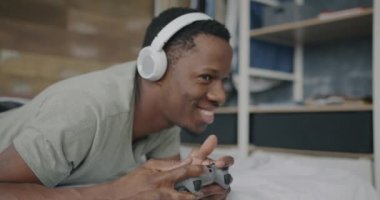Neşeli Afro-Amerikalı adam evde yatakta boş vakitlerin tadını çıkarırken joystick ve kulaklıkla video oyunu oynuyor. Oyun ve eğlence konsepti.