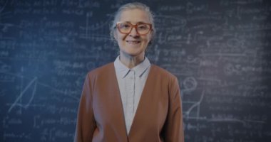 Yaşlı kadın bilim adamının yavaş çekimde karatahta duvarına yaslanmış formülle kameraya gülümseyen portresi. Eğitim ve araştırma kavramı.