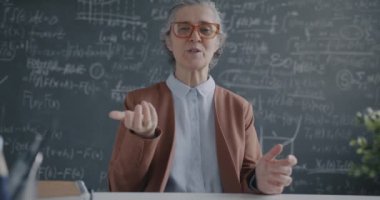 İnternette öğretmenlik yapan deneyimli matematik öğretmeninin portresi yazı tahtasının yanındaki masada oturan kameraya bakıyor. Eğitim ve sanal ders kavramı.