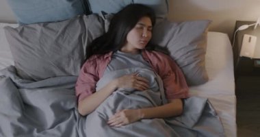 Uykulu Asyalı bir kadının yatakta uyanıp, akıllı telefonları kontrol edip karanlık bir yatak odasında uyanmasının en iyi görüntüsü. Alarm saati ve günlük rutin konsept.