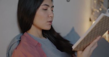 Güzel Asyalı kadın kitap okuyor. Sayfayı çevirip gülümsüyor. Akşamları rahat bir yatakta edebiyatın keyfini çıkarıyor. Hobi ve zeki insan konsepti.