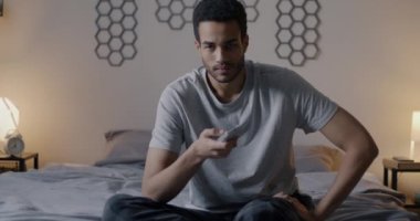 Uzaktan kumandayla televizyon izleyen Afro-Amerikan bir adamın portresi. Akşamları yatakta kameraya bakıyor. Kitle iletişim araçları ve modern yaşam tarzı kavramı.