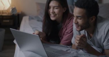Çift ırklı mutlu çift dizüstü bilgisayarla çevrimiçi video görüşmesi yapıyor ve akşamları yatakta el sallıyorlar. Aile ve iletişim konsepti.