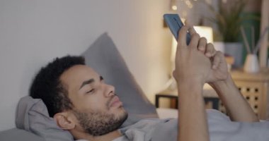 Akşamları yatakta uzanan akıllı telefonuyla sosyal medyayı kontrol eden Afro-Amerikalı bir adam. Modern teknoloji ve gençlik yaşam tarzı kavramı.