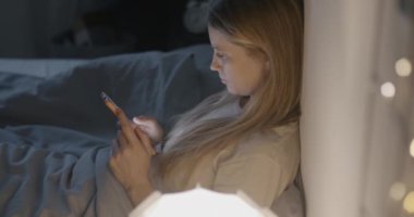 Genç bayan, uykusuz bir gecede karanlık bir yatak odasında oturmuş akıllı telefon mesajları kullanıyor. Modern teknoloji ve çevrimiçi iletişim kavramı.