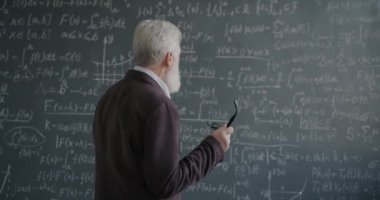 Düşünceli kıdemli profesör formüllü karatahtaya bakıyor elinde gözlüklerle üniversitede bilimi düşünüyor. Eğitim ve bilgi kavramı.