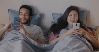 Kız ve erkek mutlu çift akıllı telefonlar kullanıyor ve sohbet ediyorlar akşamları yatakta uzanıyorlar. İlişki ve aygıt kavramı.