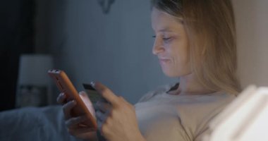 Akıllı telefon kullanan ve elinde kredi kartı tutan mutlu genç bir kadın akşamları yatakta alışveriş yapıyor. E-ticaret ve para kavramı.