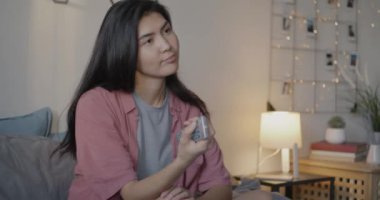 Genç Asyalı bayan televizyonda uzaktan kumandayla eğlenceli bir program izliyor. Yatak odasında tek başına eğleniyor. Kitle iletişim araçları ve boş zaman etkinliği kavramı.