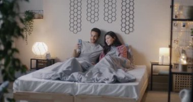 Çift ırklı mutlu erkek ve kadın akıllı telefonlarıyla online video görüşmesi yapıyor ve akşamları yatakta oturup gülüyor. İletişim ve ilişki kavramı.
