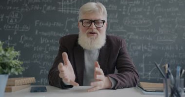 Son sınıf profesörünün portresi fen bilgisi öğretiyor ve sınıftaki masada oturan kameraya bakarak el kol hareketi yapıyor. Sanal ders ve eğitim kavramı.