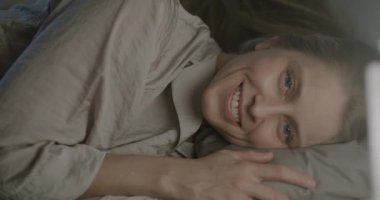 Yatakta, yumuşak yastığa uzanmış kameraya gülümseyen güzel bir genç kadının portresi. Pozitif duygu ve uyku zamanı konsepti.