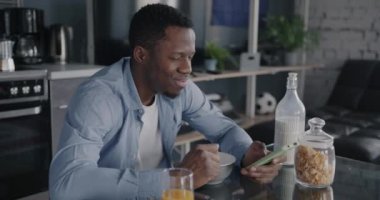 Afro-Amerikalı neşeli adam sağlıklı kahvaltı yapıyor ve apartmandaki sosyal medyada akıllı telefon mesajları kullanıyor. Modern cihaz ve yaşam tarzı konsepti.