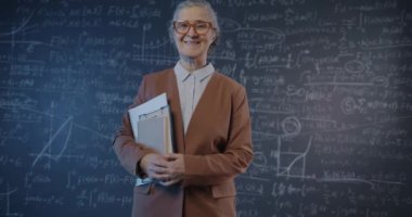 Üniversite sınıfında elinde kitaplarla gülümseyen karatahta duvarlı yaşlı bayan profesörün yavaş çekimde portresi. Eğitim ve araştırma kavramı.