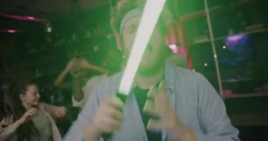 Neon lamba kılıcıyla dans eden neşeli adamın yavaş çekimde portresi. Akşamları apartmanda arkadaşlarıyla eğleniyor. Parti ve gençlik yaşam tarzı kavramı.