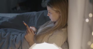 Genç bayan, uykusuz bir gecede karanlık bir yatak odasında oturmuş akıllı telefon mesajları kullanıyor. Modern teknoloji ve çevrimiçi iletişim kavramı.